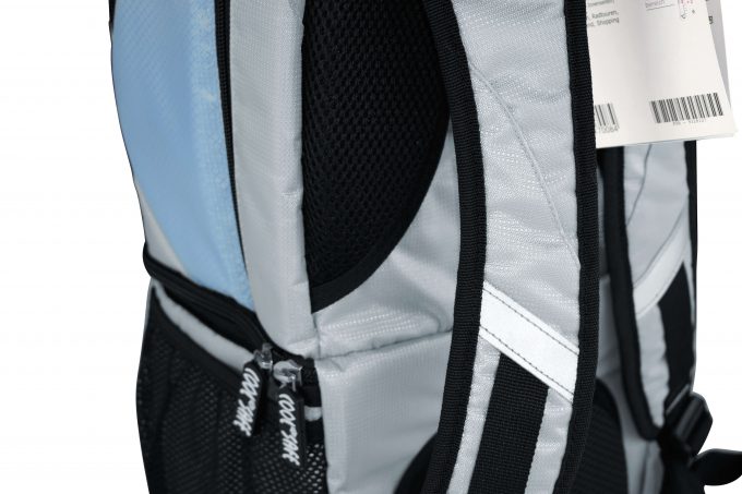 Seitenansicht des silbergrauen COOL*SAFE-Rucksacks mit schwarzer Netz-Seitentasche, reflektierendem Streifen und gepolstertem Rückenbereich. Ein Produktanhänger ist am Tragegurt befestigt.