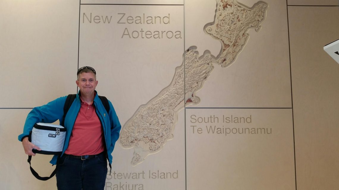 Person mit Rucksack auf dem Rücken und einer COOL*SAFE Kühltasche vor einer Wandkarte von Neuseeland mit den Bezeichnungen Aotearoa, South Island und Stewart Island.
