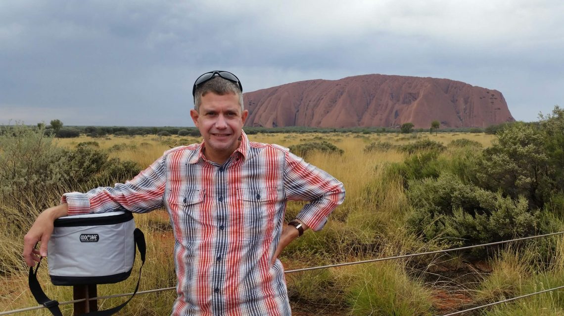 Mann mit kariertem Hemd lehnt an einer weißen COOL*SAFE Kühltasche, im Hintergrund Uluru in der australischen Wüstenlandschaft.