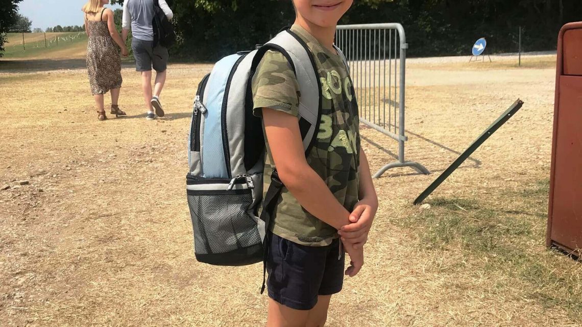 Kind mit Sonnenbrille, Cap und Shorts trägt eine COOL*SAFE Medikamentenkühltasche als Rucksack auf einem trockenen Sommerfeld.