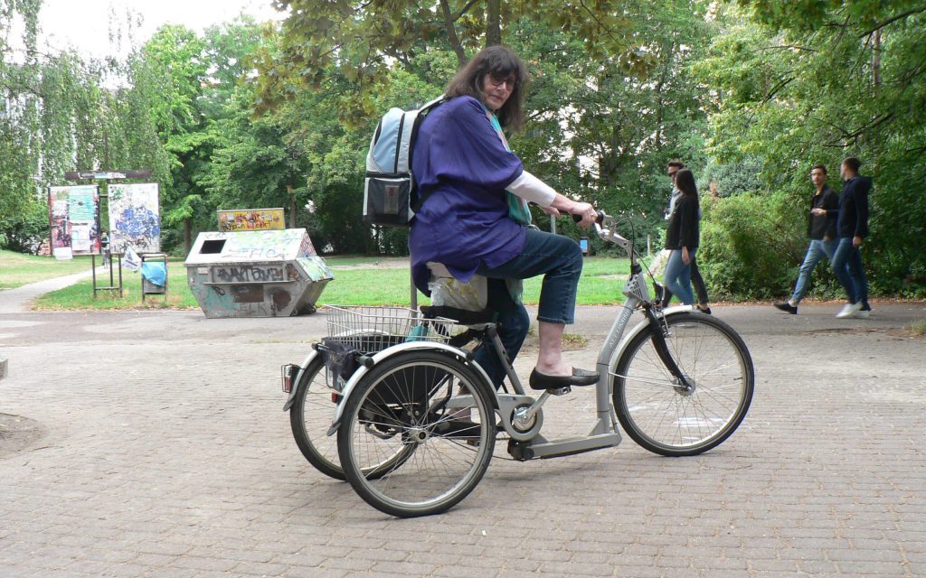 Frau mit COOL*SAFE Rucksack fährt auf einem Dreirad durch einen Park, im Hintergrund Bäume und Passanten.