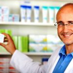 dreamstime_xxl_31328989-Apotheker-COOLSAFE Lächelnder Apotheker mit Brille und Apothekerkittel zeigt eine Medikamentenpackung in einer Apotheke vor einem Regal mit Arzneimitteln.