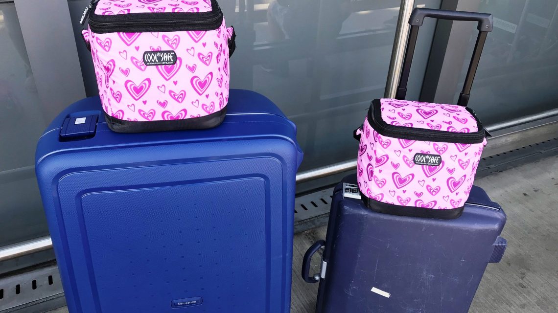 Zwei pinkfarbene COOL*SAFE Medikamentenkühltaschen mit Herzmuster stehen auf zwei dunkelblauen Rollkoffern vor einem Flughafengebäude.