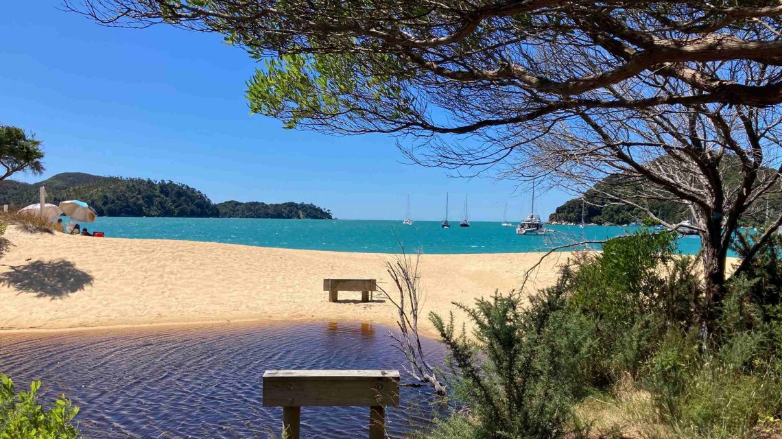 Blick durch Bäume auf einen goldenen Sandstrand mit zwei Sitzbänken, türkisblauem Meer und Segelbooten in einer Bucht.