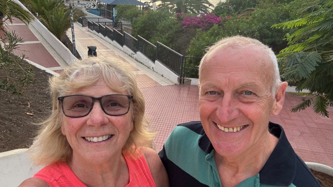 Lächelndes älteres Paar vor mediterraner Kulisse mit Palmen, Blüten und Spazierweg bei Sonnenuntergang.