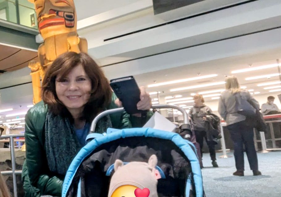 Frau mit Kinderwagen in einem Flughafenbereich, im Hintergrund ein Totempfahl und wartende Reisende. Das Kind im Wagen ist durch ein Emoji mit Herzchen unkenntlich gemacht.