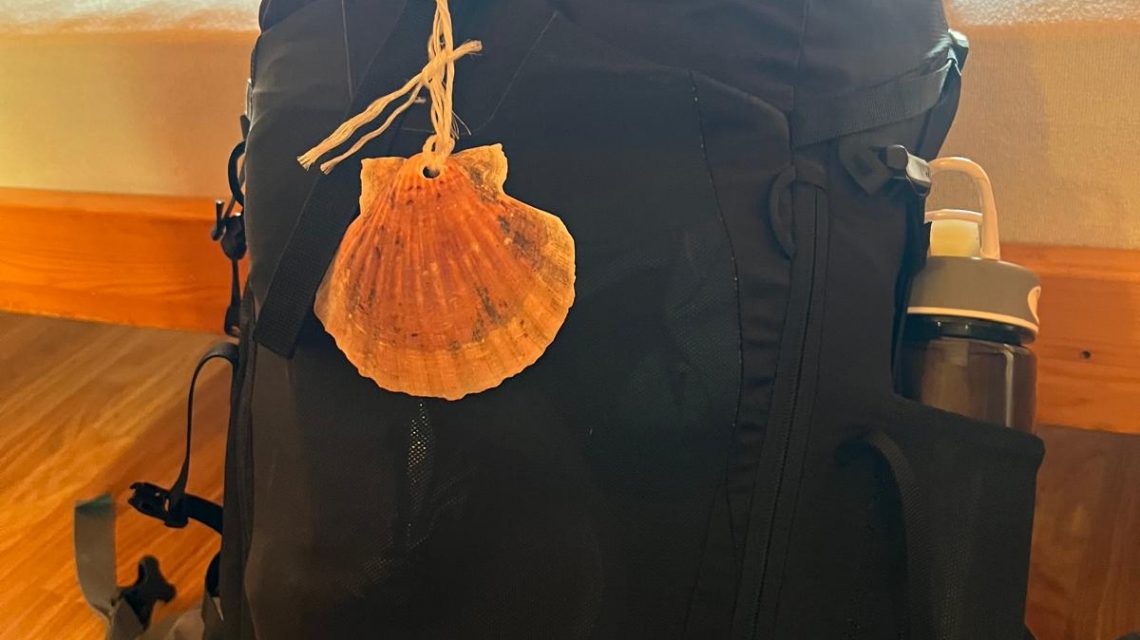 Schwarzer Wanderrucksack mit einer Pilgermuschel als Erkennungszeichen auf dem Jakobsweg, seitlich befestigt eine COOL*SAFE Medikamentenkühltasche.